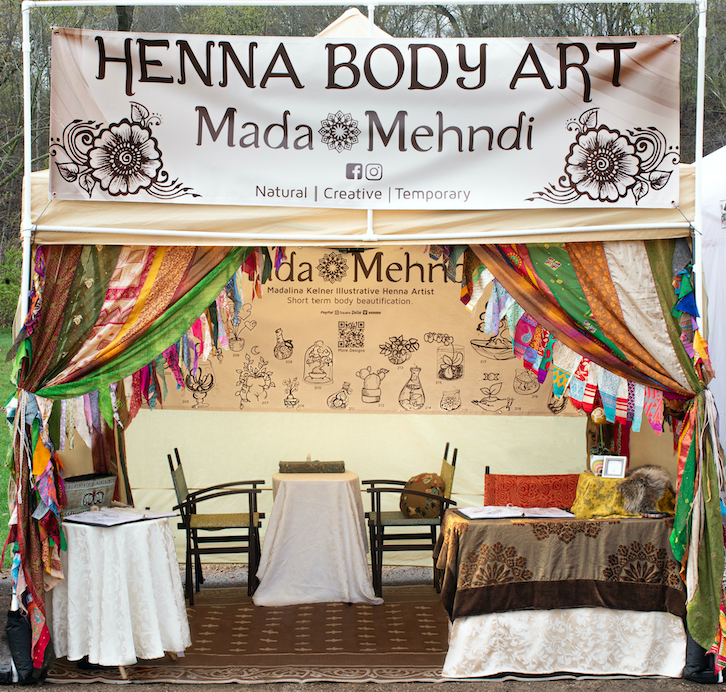 madalinas henna booth. one table at the front. tan tent and cozy curtons making the space warm and inviting. there is a table and chairs set up in the back where the henna artist takes apointments.