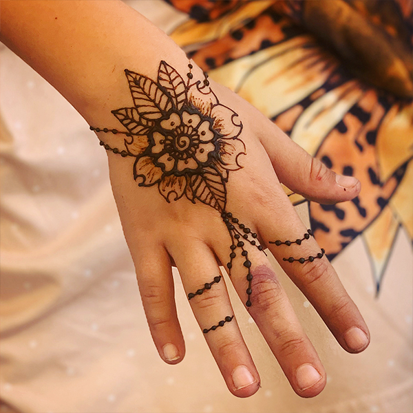 Medium skin tone hand of young individual over a blurred background of a flower. The henna design of a flower bracelet with beadwork draping over the fingers.